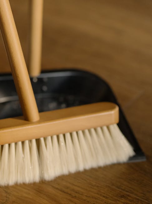 A close-up of a wooden-handled broom with beige bristles, positioned on a dark wooden floor in a room with warm lighting. The broom is resting inside a black dustpan, which is visible in the backgroun
