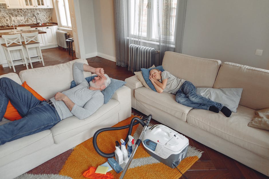 A young woman wearing a lavender sweatshirt, yellow rubber gloves, and checkered pants is kneeling on the dark hardwood floor of a modern living room, wiping a small round glass coffee table with a pi